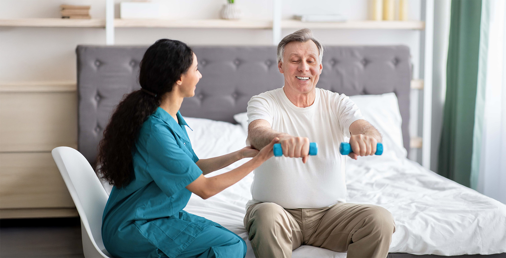 Young physiotherapist helping homebound elderly man to do physical exercises with dumbbells on bed at home